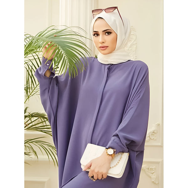 Une femme portant une longue robe violette à manches bouffantes et un hijab blanc. Elle porte des lunettes de soleil et un sac à main blanc.