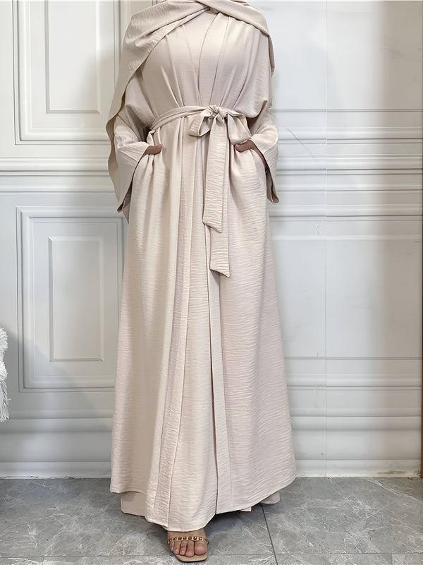 Une femme portant une longue robe beige avec une ceinture à la taille et une écharpe assortie. La robe est ample et fluide, avec des manches longues et un col montant. La femme porte des sandales beige.