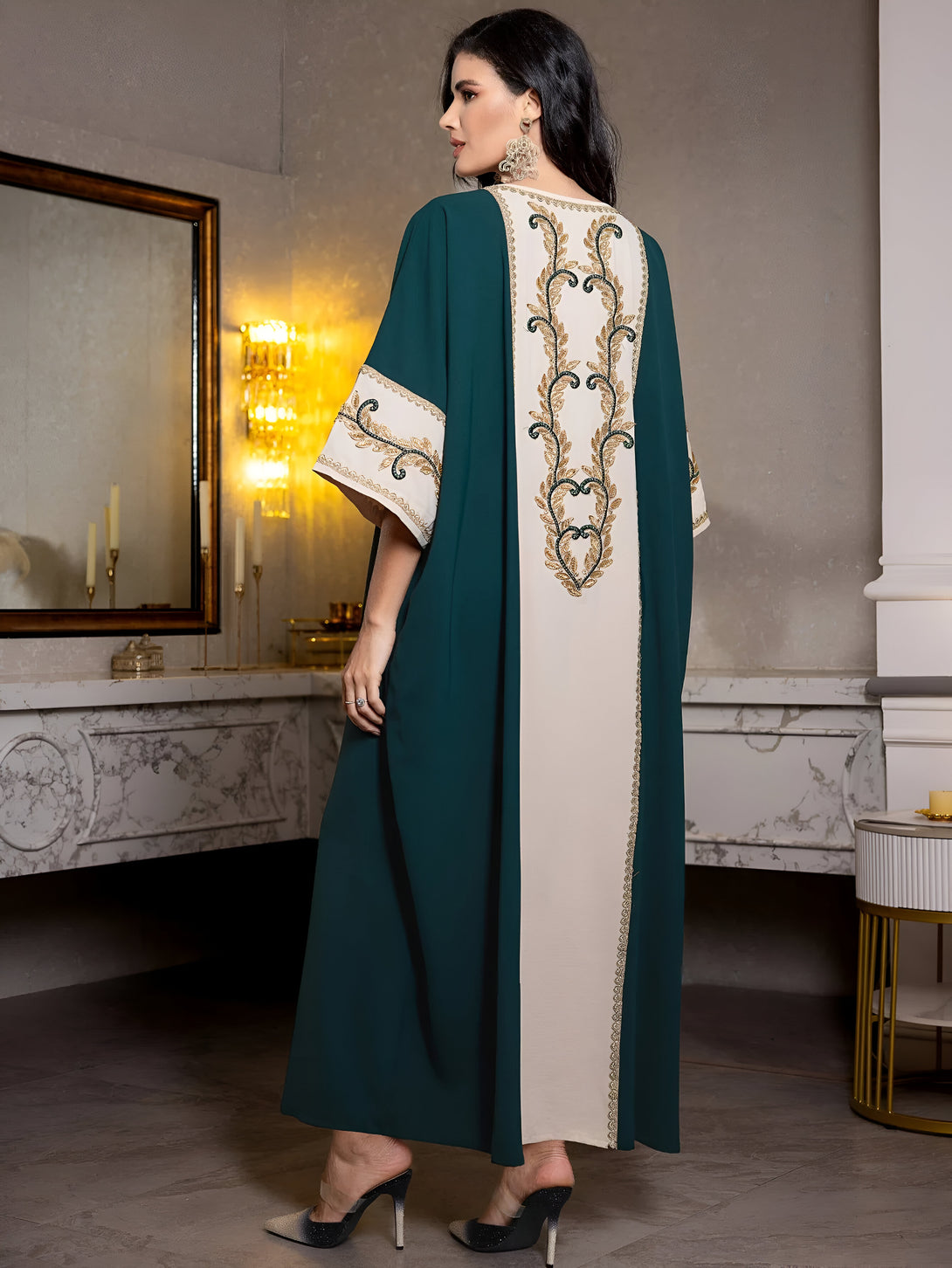 Une femme portant une abaya verte et blanche avec des broderies dorées sur le dos. La robe est à manches trois-quarts et a une coupe ample. La femme porte des talons hauts.