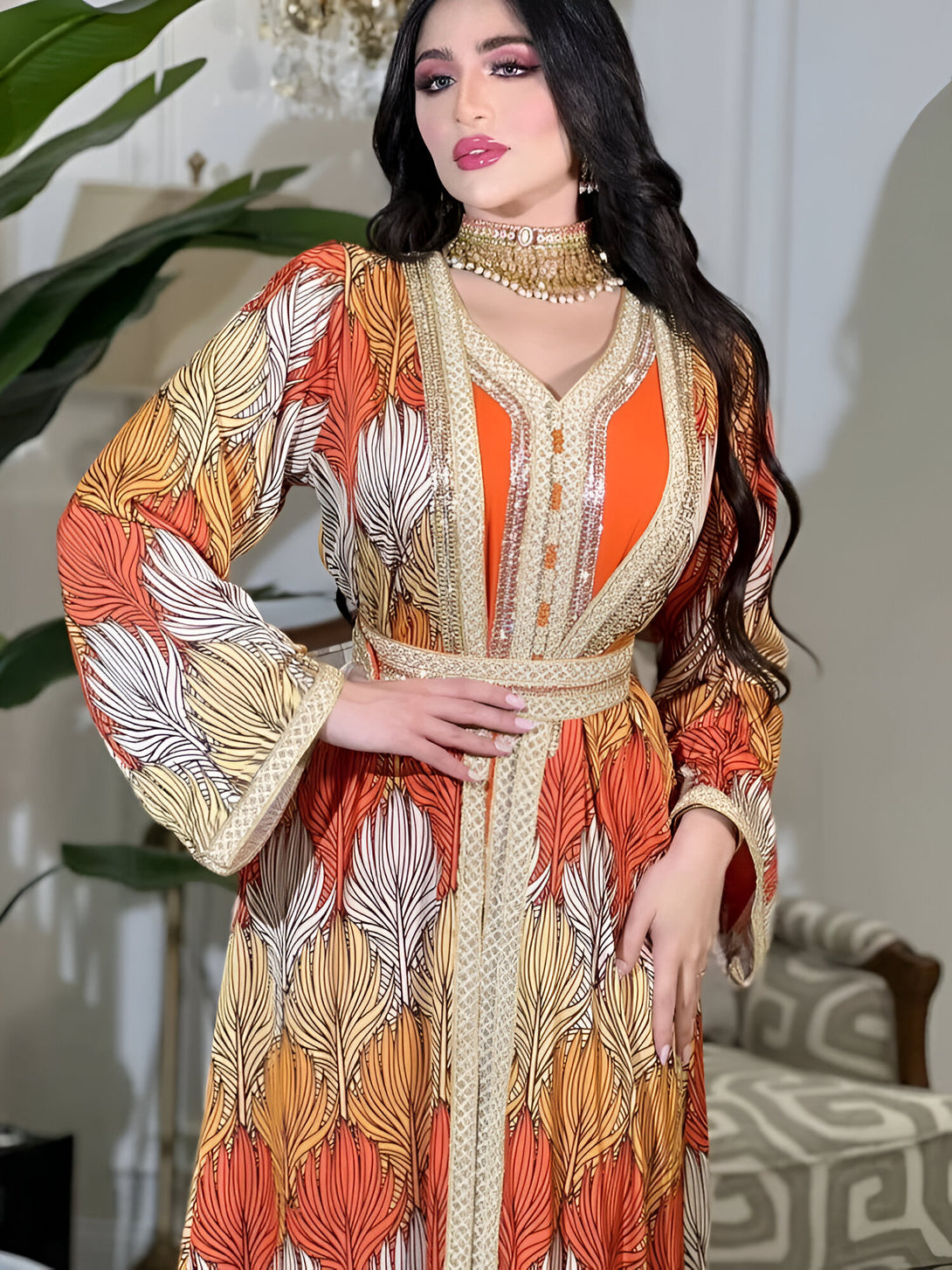 Une femme élégante porte une robe longue de style marocain avec des motifs orange, beige et blancs. La robe est ornée d'une bordure dorée et d'une ceinture.
