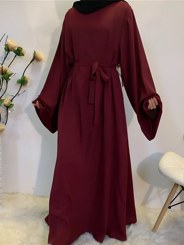 Une robe longue de couleur bordeaux avec une ceinture assortie, idéale pour les femmes musulmanes.  La robe a des manches longues et une coupe ample.