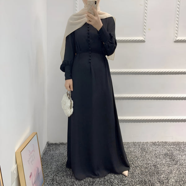 Une femme portant une longue robe noire boutonnée avec un hijab beige. Elle tient un sac à main blanc et pose devant un mur blanc.