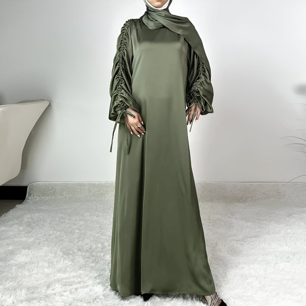 Une femme porte une robe longue verte en satin avec des manches bouffantes et un hijab assorti. La robe est ample et fluide, idéale pour les occasions spéciales.