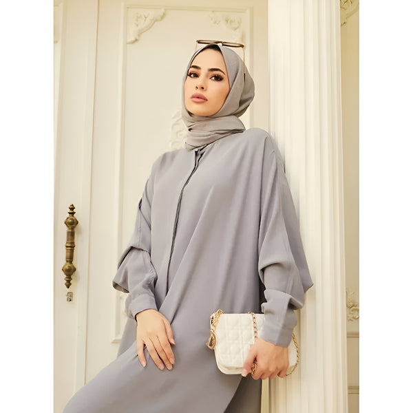 Une femme portant une robe grise à manches longues et un hijab gris, tenant un sac à main blanc.