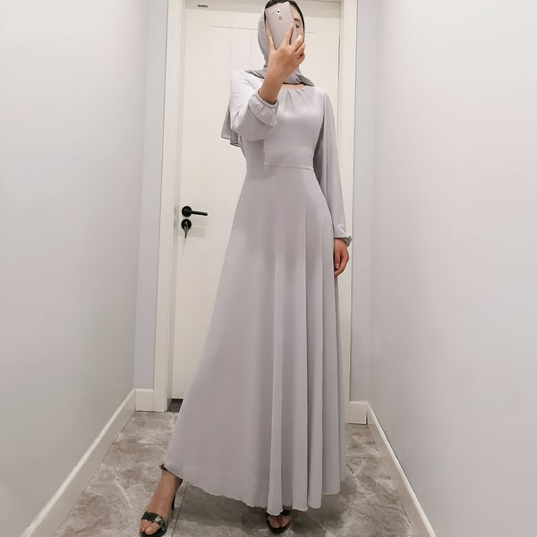 Une femme porte une longue robe grise à manches longues avec un hijab assorti. La robe a une coupe ample et fluide, idéale pour les occasions spéciales.