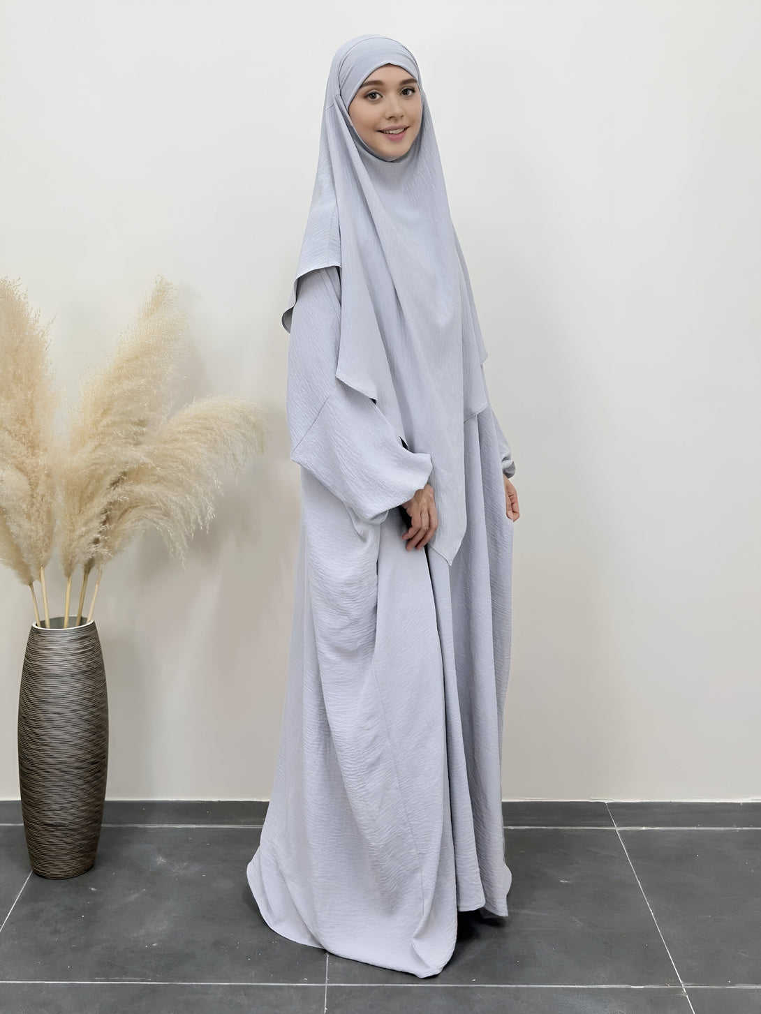 Une femme portant une abaya et un hijab gris clair, souriante et élégante. La robe est ample et fluide, parfaite pour une occasion spéciale.