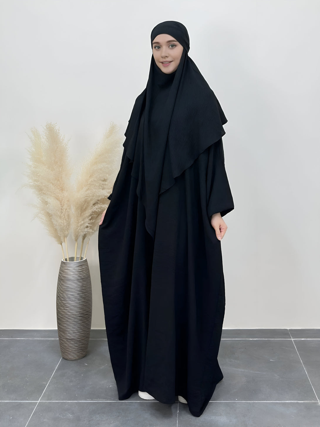 Une femme portant une abaya noire ample avec un hijab assorti, debout devant un mur blanc et un vase avec des fleurs séchées.