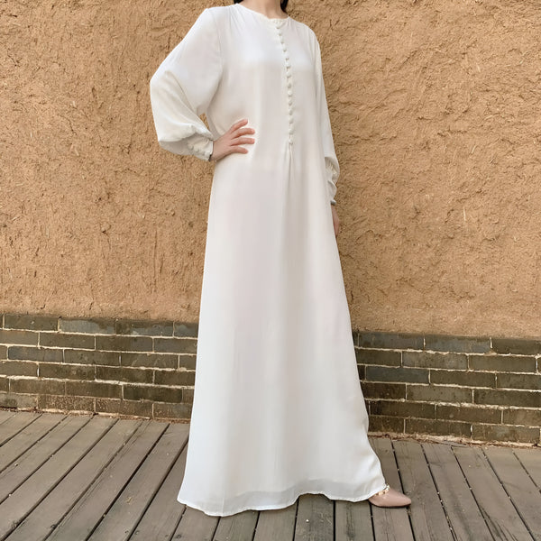 Une robe longue blanche boutonnée à l'avant avec des manches bouffantes, portée par une femme sur un fond en brique.