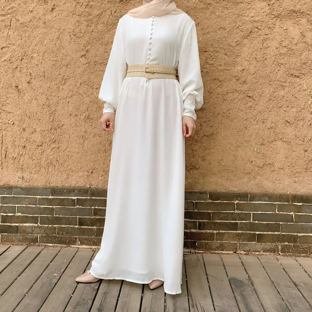 Une femme porte une robe blanche longue avec une ceinture beige, debout devant un mur de briques.