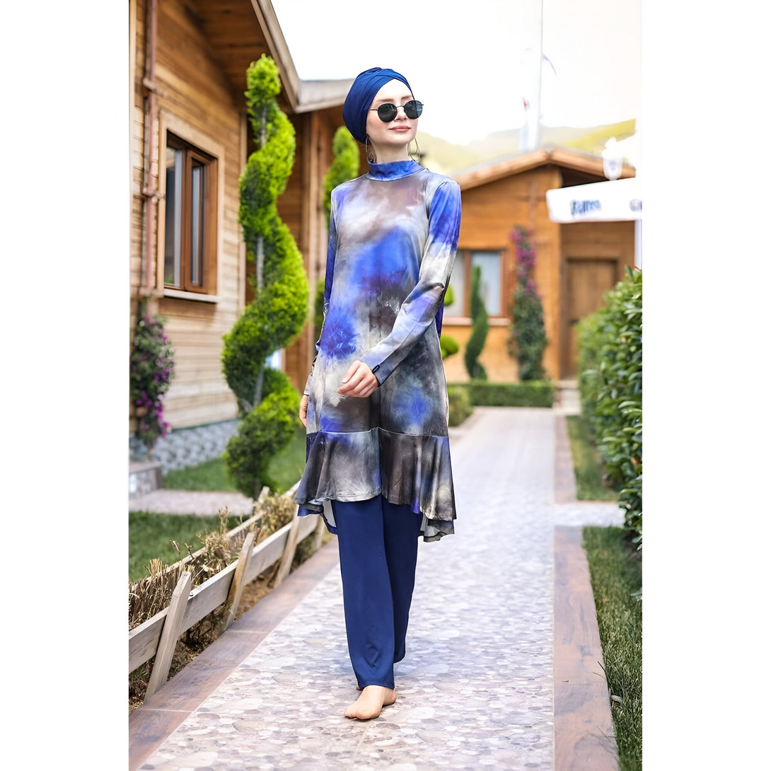 Une femme portant un hijab et une tenue de bain musulmane bleue et grise à motifs. Elle porte des lunettes de soleil et marche sur un chemin pavé devant un bâtiment en bois.