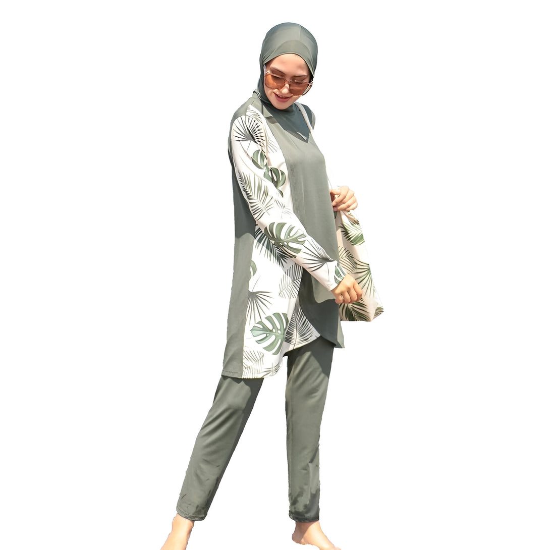 Une femme portant un maillot de bain musulman vert avec un motif de feuilles tropicales.  Le maillot de bain est composé d'une tunique à manches longues et d'un pantalon long. La femme porte également un hijab vert.