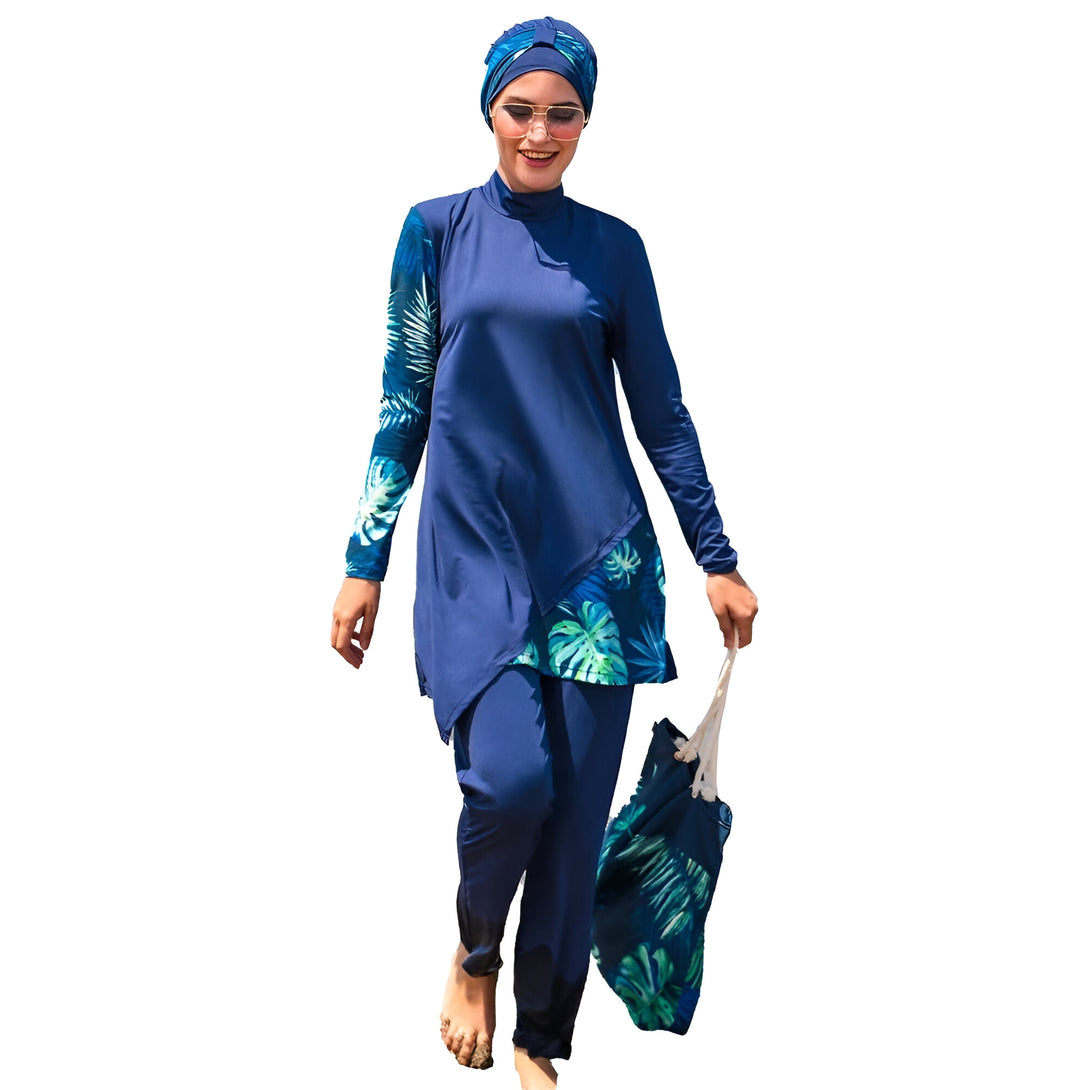 Une femme portant un hijab et un maillot de bain musulman bleu foncé avec un imprimé tropical vert. Elle tient un sac en toile blanc dans sa main droite et marche pieds nus sur la plage.