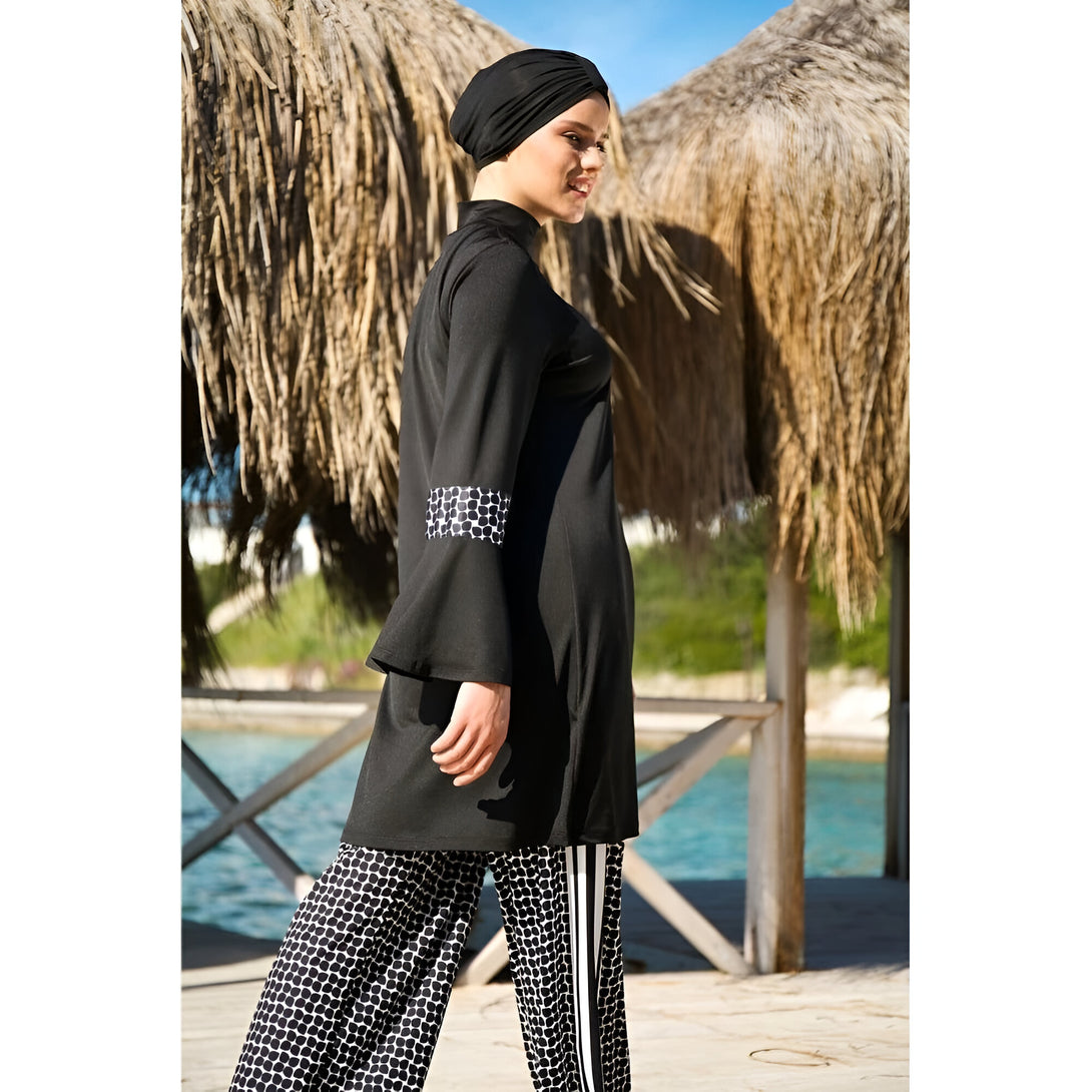 Une femme portant un hijab noir et un maillot de bain musulman noir et blanc à motifs géométriques. Elle se tient devant un palmier sur une plage.