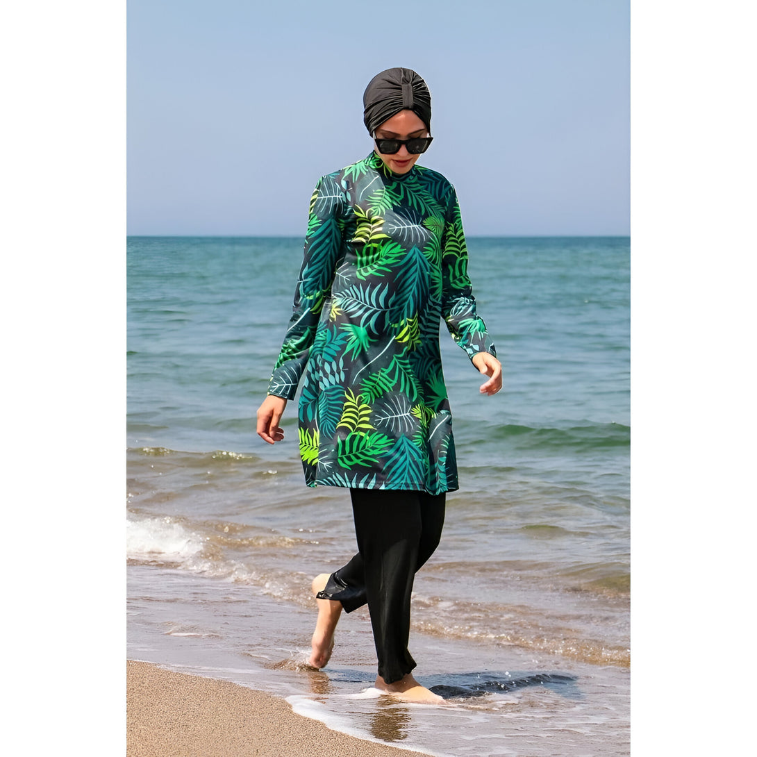 Une femme portant un maillot de bain musulman avec un motif de feuilles vertes sur fond bleu foncé, marchant sur la plage.