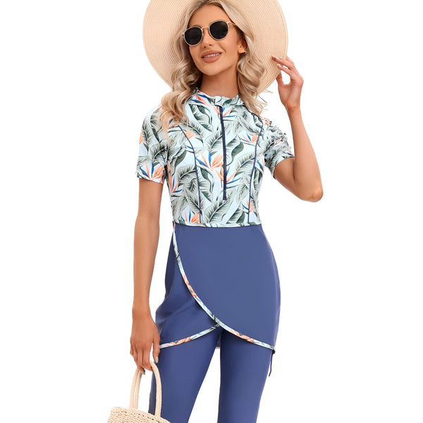 Une femme portant un maillot de bain musulman à manches courtes avec un motif tropical. Le maillot de bain est bleu marine et bleu clair avec une jupe-culotte intégrée. Elle porte également un chapeau de paille et des lunettes de soleil.