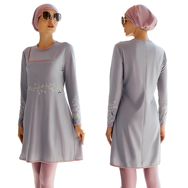 Une femme porte un maillot de bain musulman gris avec des détails floraux brodés. Le maillot de bain est à manches longues et a une jupe intégrée. La femme porte également un foulard rose et des lunettes de soleil.