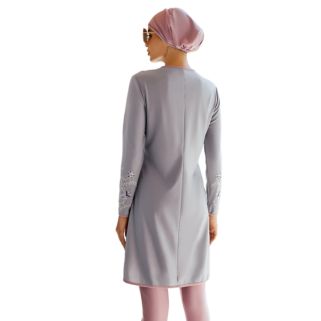 Une femme portant un maillot de bain musulman gris avec des détails floraux. Le maillot de bain est à manches longues et couvre les jambes. La femme porte également un foulard rose.