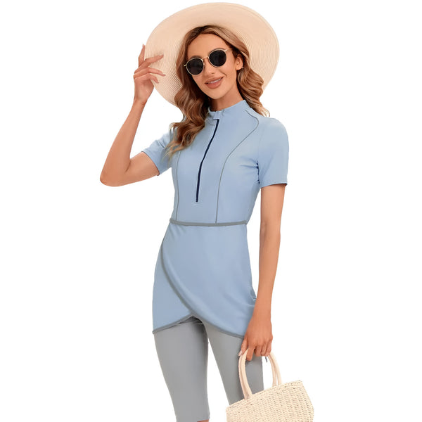 Une femme portant un maillot de bain islamique bleu clair avec un col montant et une fermeture éclair. Elle porte également un chapeau de paille et des lunettes de soleil.