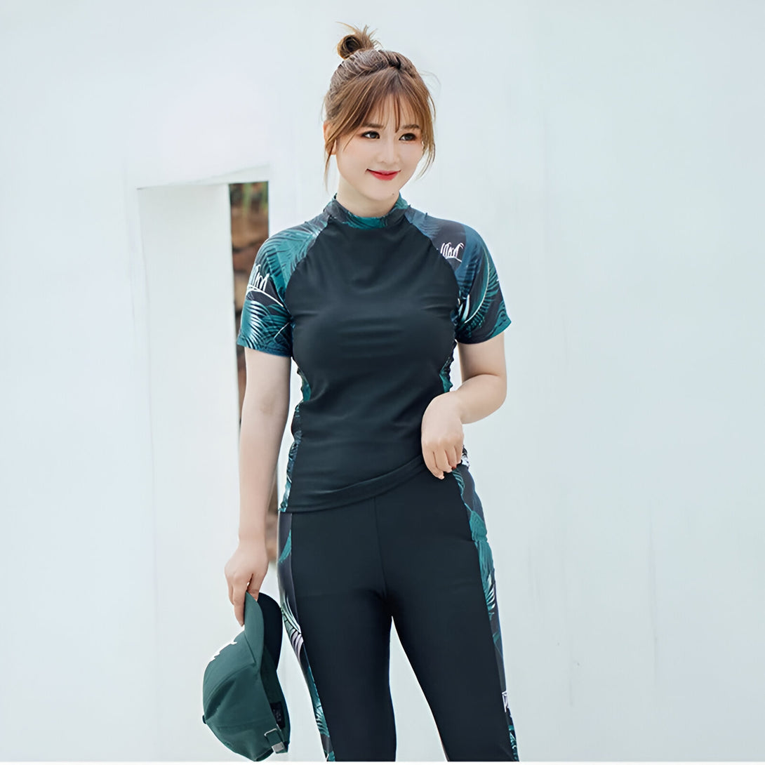 Une femme portant un maillot de bain musulman noir et vert à manches courtes avec un imprimé de feuilles. Elle porte également un pantalon noir et un chapeau vert.