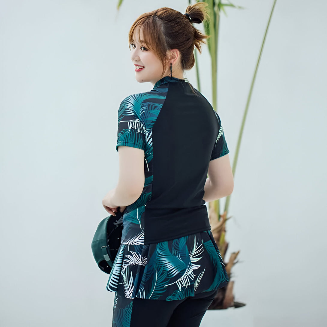 Une femme portant un maillot de bain musulman avec un motif de feuilles tropicales. Le maillot de bain est de couleur noire et verte avec des manches courtes et une jupe courte.