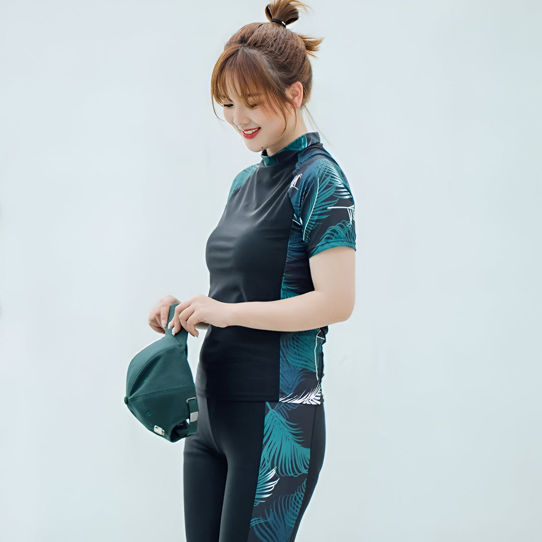 Une femme porte un maillot de bain musulman à manches courtes noir avec un motif de feuilles vertes. Elle porte également un pantalon de bain assorti. Elle tient une casquette verte dans sa main.