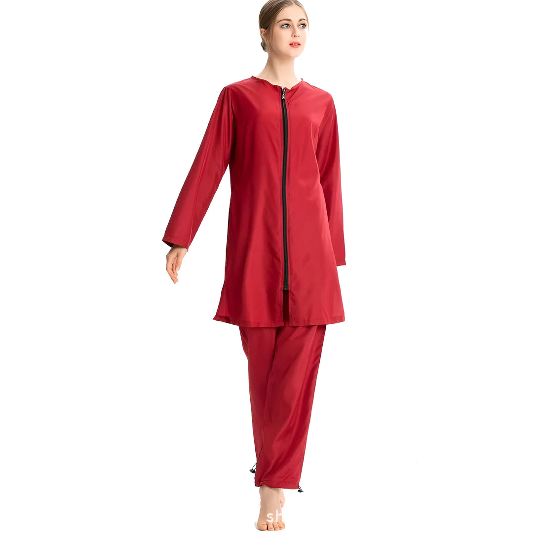 Une femme porte un maillot de bain musulman rouge à manches longues et pantalon long. Le maillot de bain est zippé sur le devant et a une coupe ample. La femme a les cheveux tirés en arrière et porte un maquillage léger. Elle pose sur fond blanc.