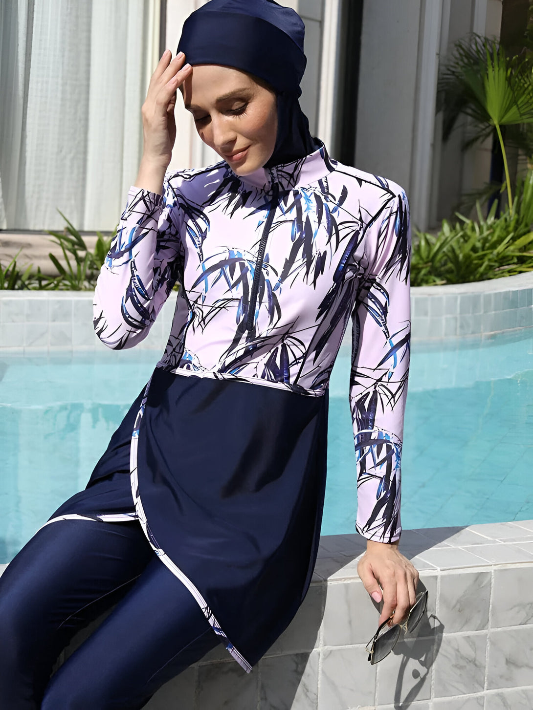 Une femme portant un maillot de bain musulman bleu et rose à motifs, avec un hijab bleu foncé, assise au bord d'une piscine.