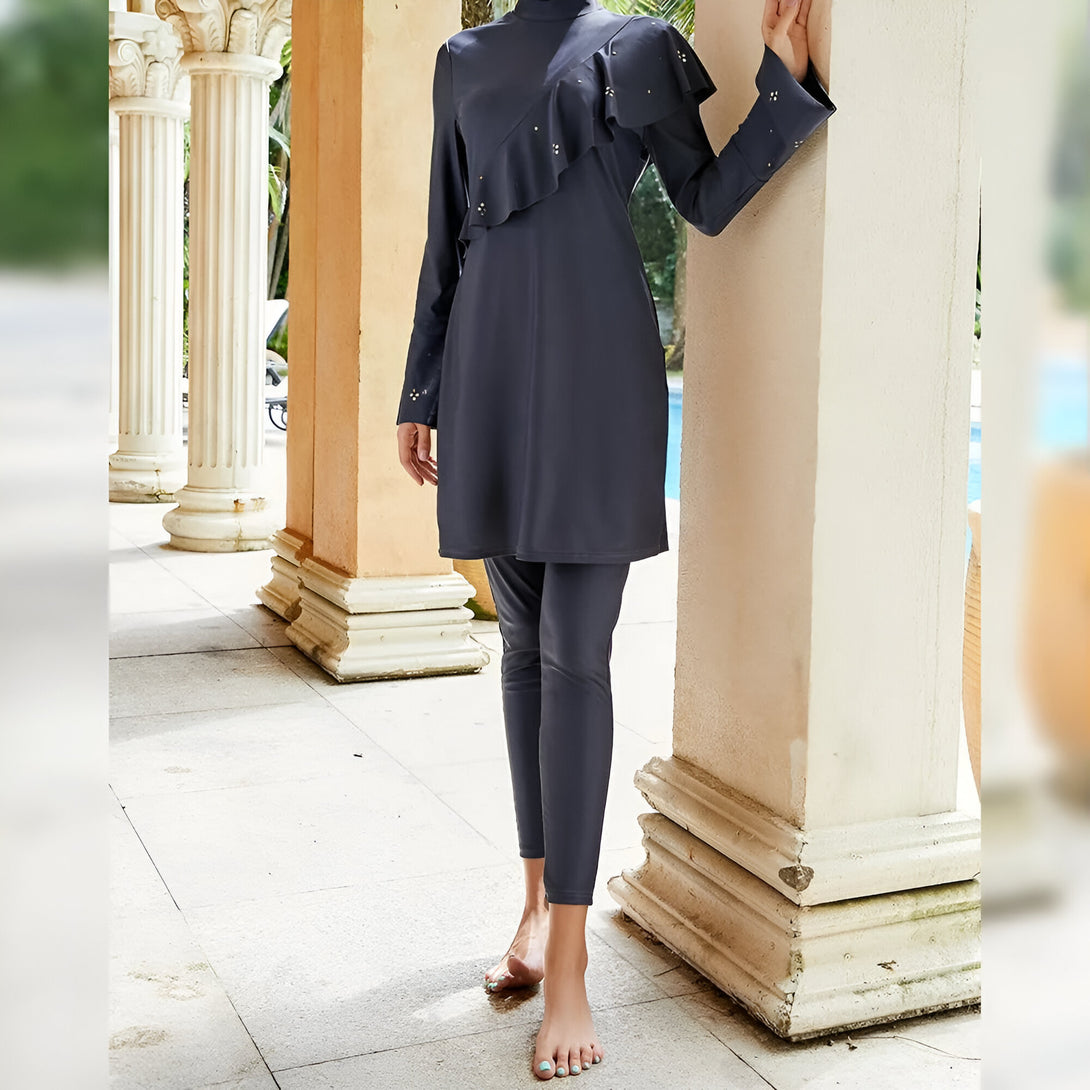 Une femme portant un maillot de bain musulman gris avec des manches longues et une jupe longue. Le maillot de bain a des volants sur le devant. La femme est debout devant des colonnes en pierre.