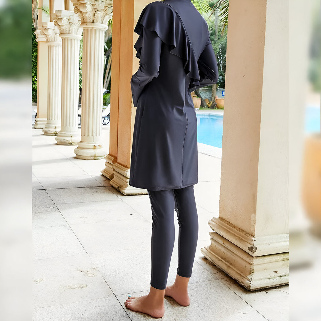 Une femme portant un maillot de bain musulman noir avec une jupe et un pantalon long. Le maillot de bain a une frise sur le devant. La femme est debout près d'une piscine.