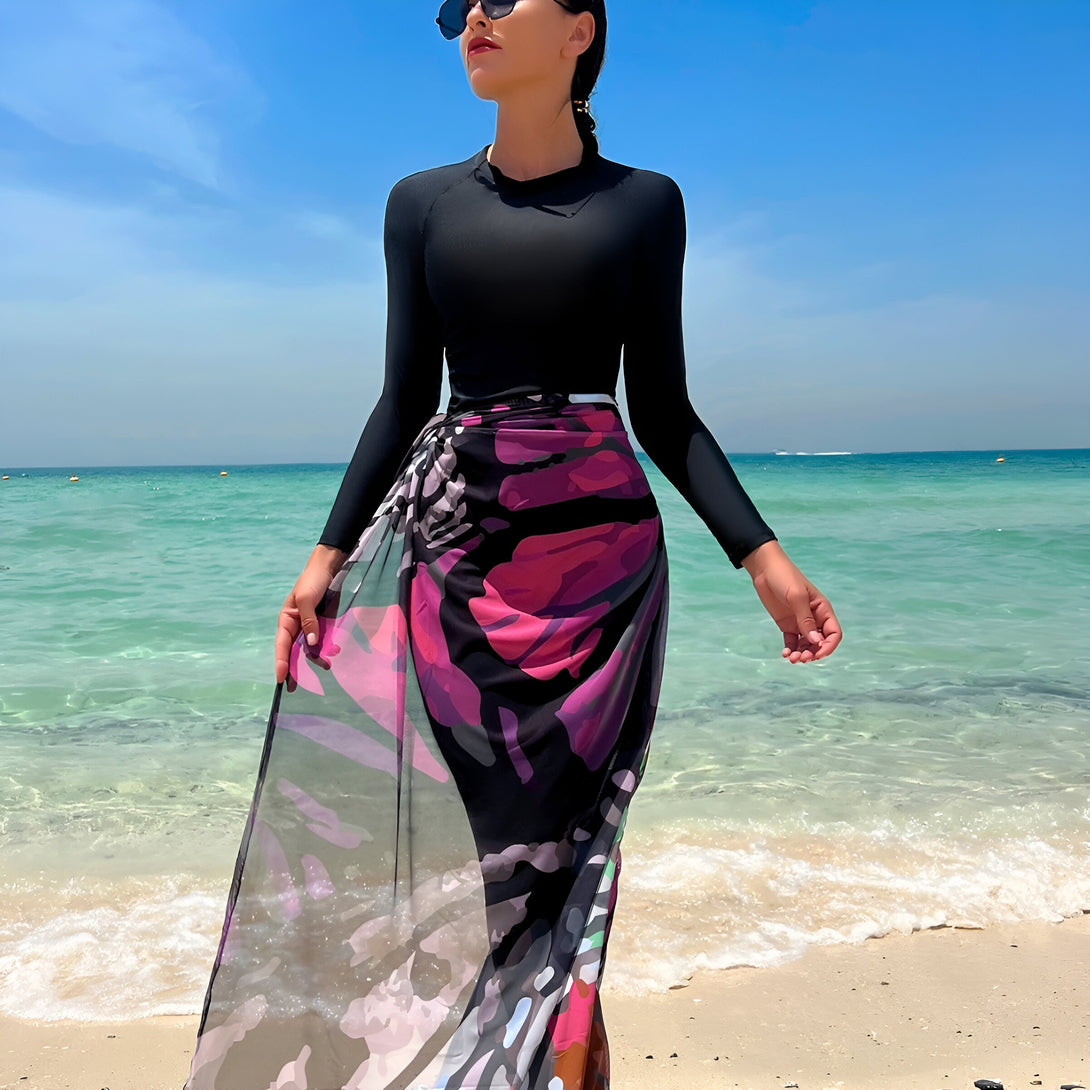 Une femme portant un maillot de bain musulman noir à manches longues et une jupe longue à imprimé floral se tient sur une plage de sable blanc. Elle porte des lunettes de soleil et regarde vers le ciel bleu.