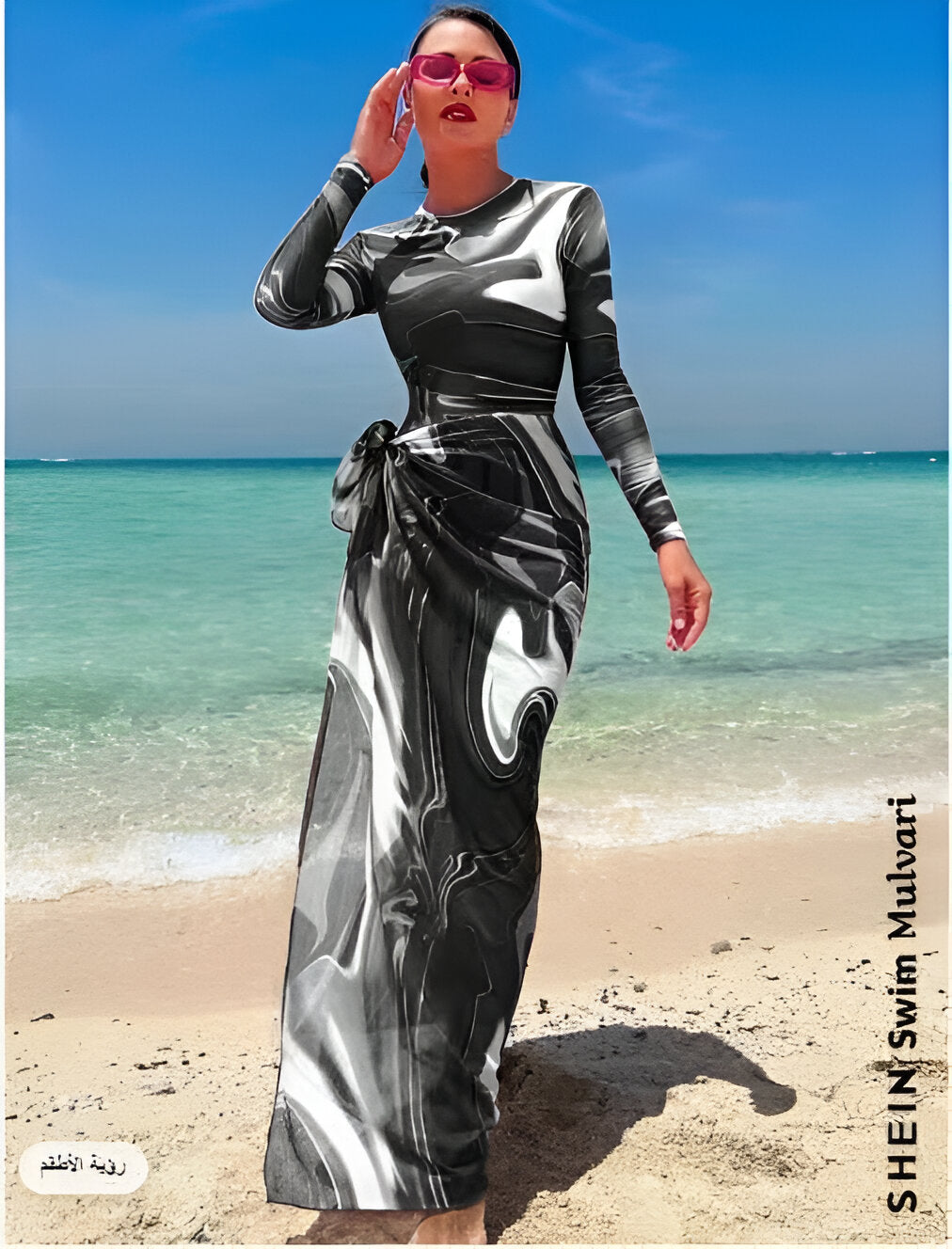 Une femme portant une robe longue noire et blanche à imprimé marbré, se tenant sur une plage de sable. La robe est élégante et flatteuse, avec une coupe moulante qui met en valeur sa silhouette.