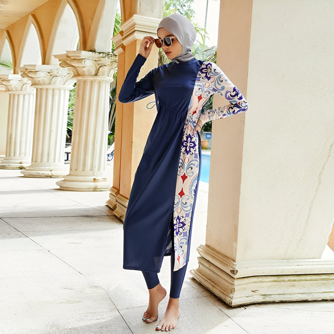 Une femme portant un maillot de bain musulman bleu foncé avec des détails imprimés. Le maillot de bain comprend une tunique à manches longues et un pantalon long. Elle porte un hijab gris et des lunettes de soleil.