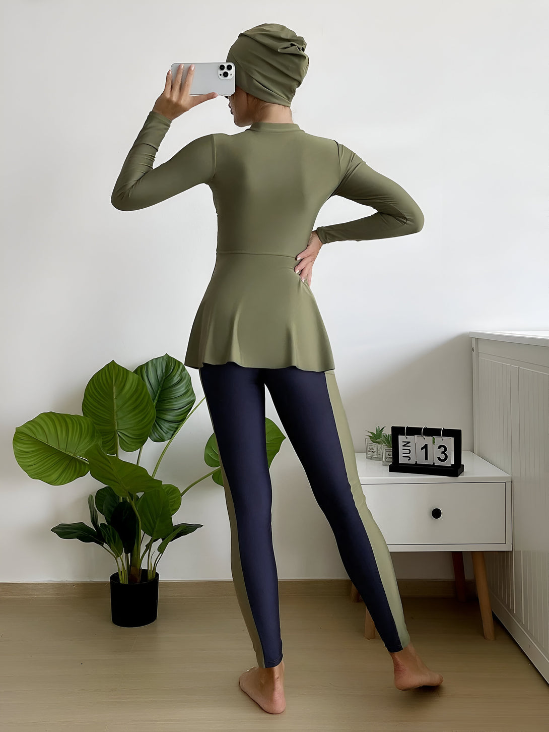 Une femme portant un hijab vert et un maillot de bain musulman vert et bleu marine. Elle est debout devant un mur blanc, avec une plante verte à côté d'elle.