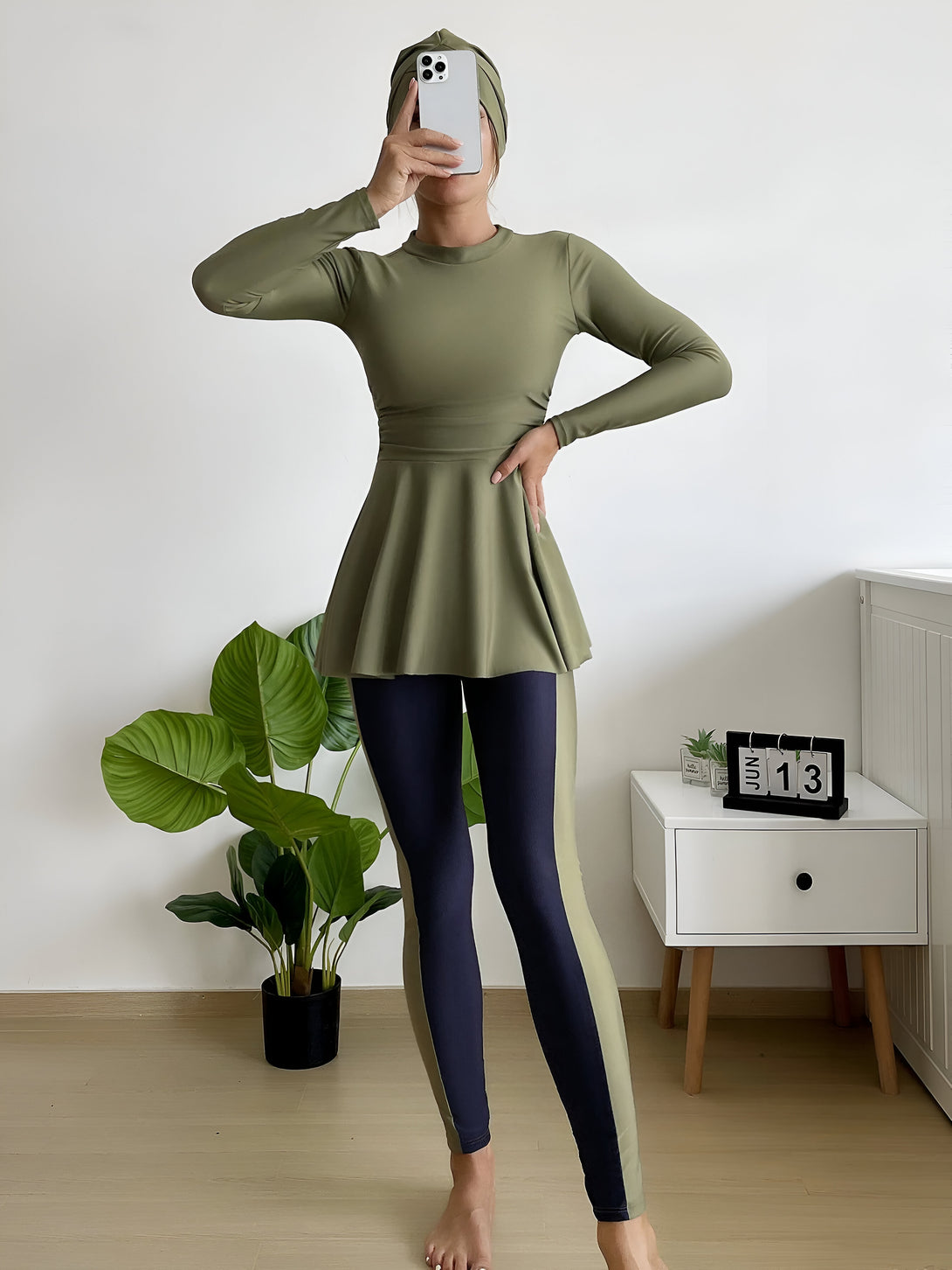 Une femme portant un maillot de bain islamique vert avec un hijab assorti et un pantalon noir. Elle est debout dans une pièce avec un plant de fond et un petit bureau.