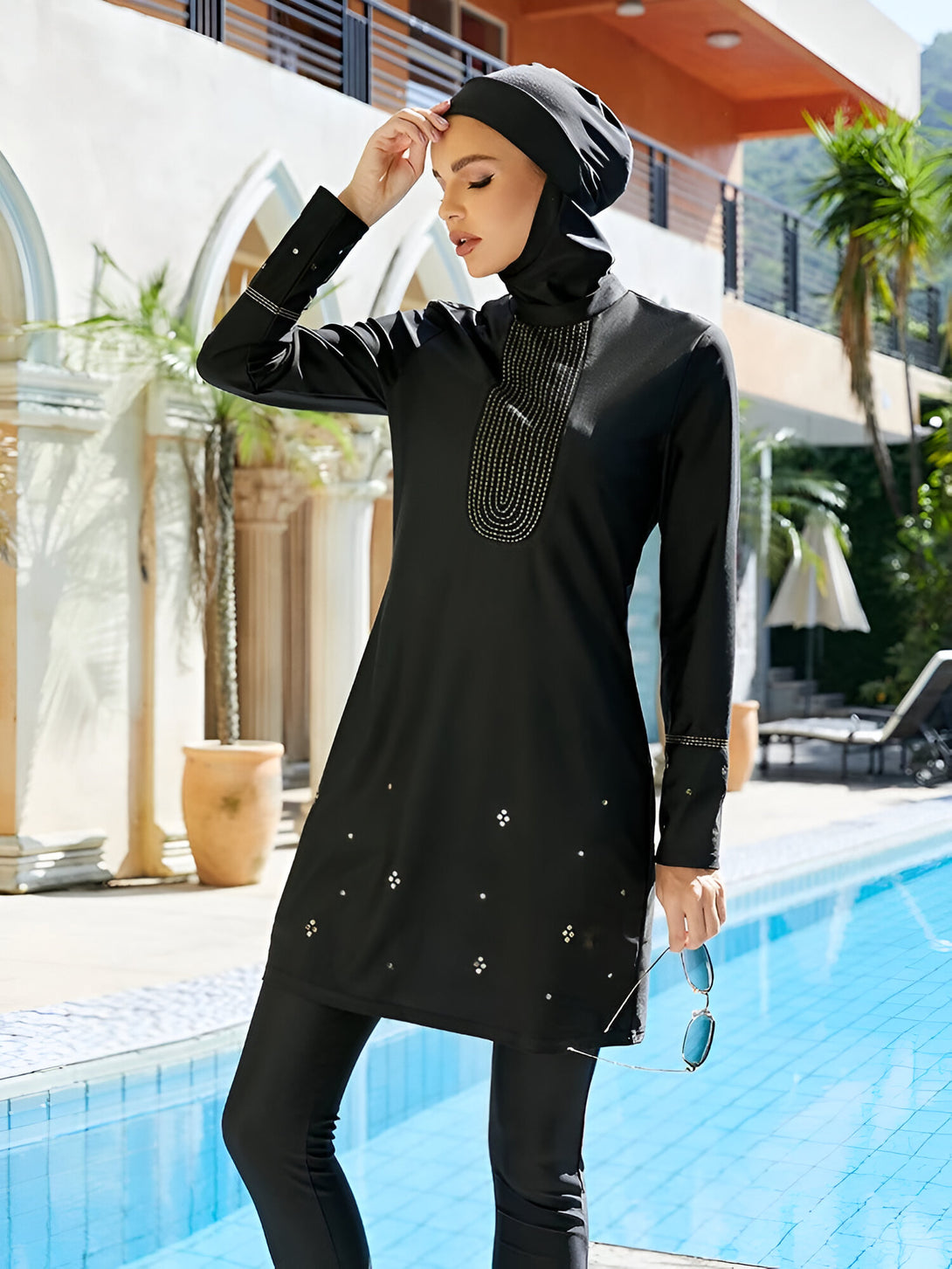 Une femme portant un maillot de bain musulman noir avec des détails dorés et des perles, près d'une piscine.