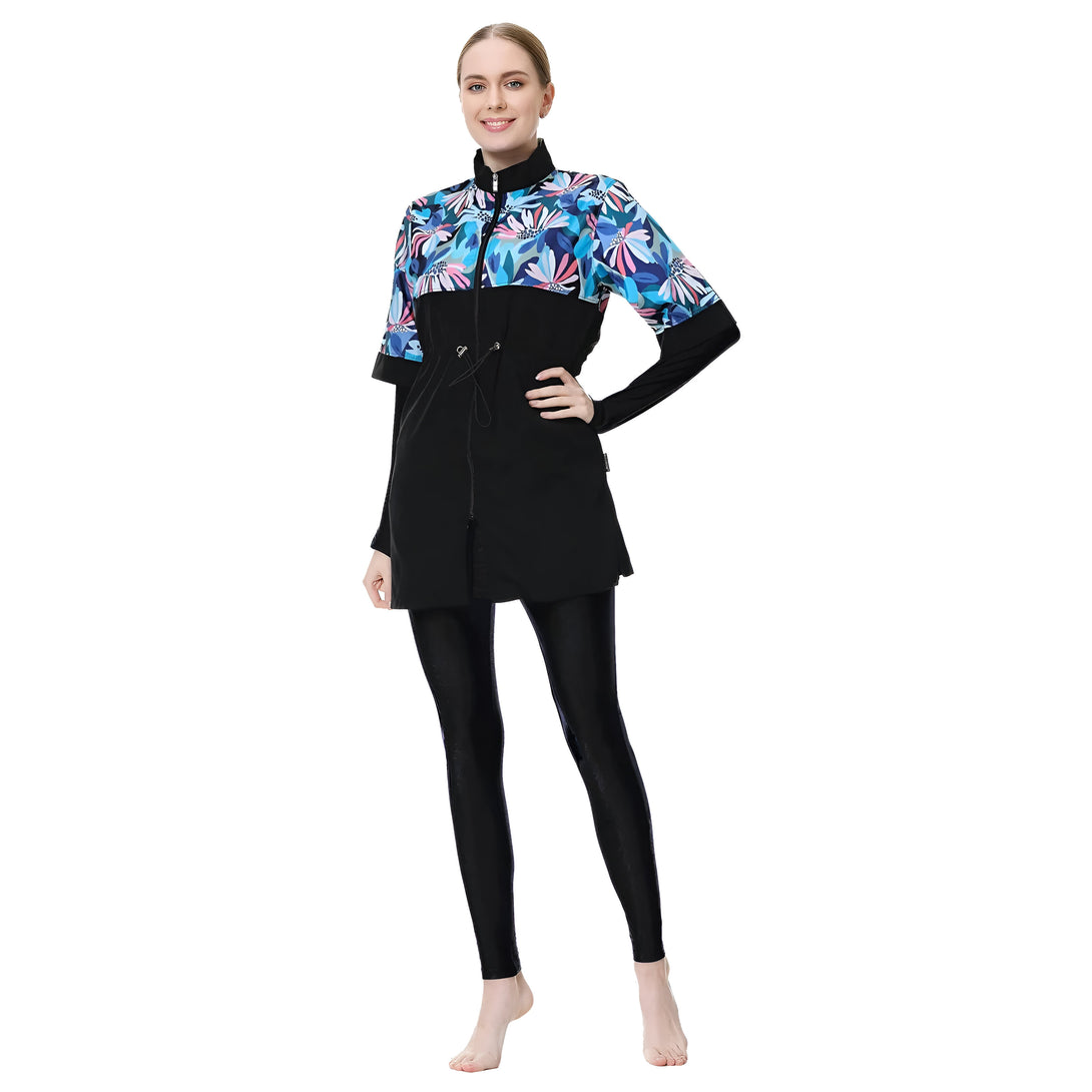 Une femme porte un maillot de bain musulman noir avec un haut à imprimé floral bleu et rose. Le maillot de bain est à manches longues et a un col montant. La femme porte également un pantalon noir.