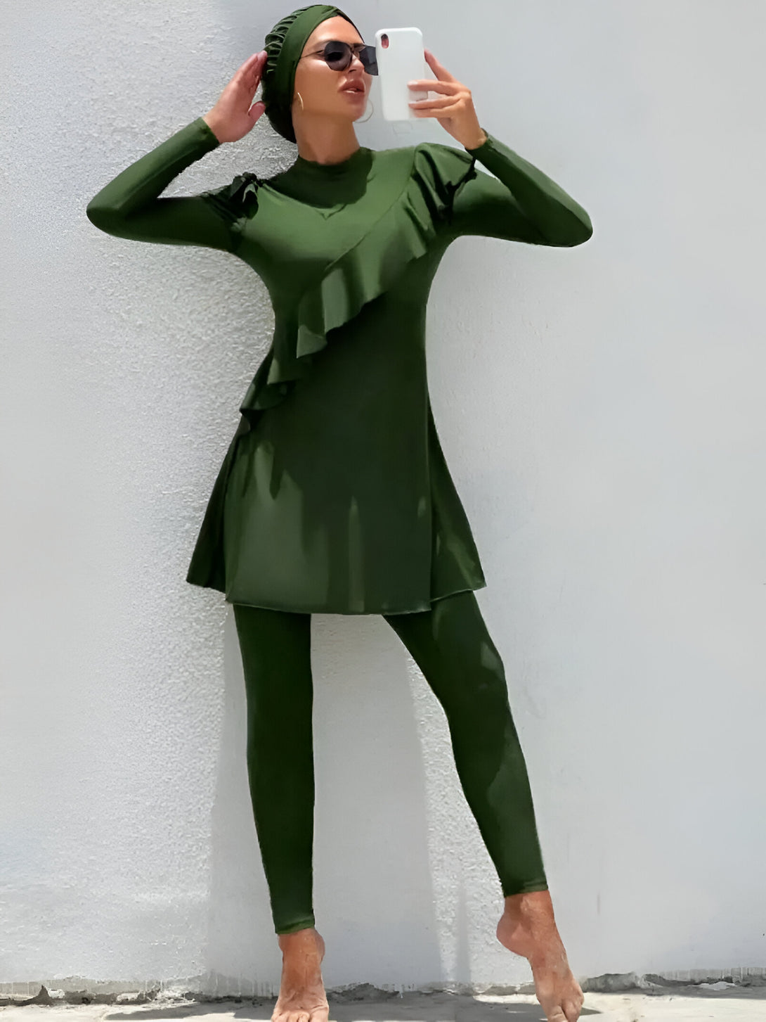 Une femme portant un maillot de bain musulman vert avec une jupe plissée et un hijab.  Le maillot de bain est à manches longues et a un pantalon long. La femme porte des lunettes de soleil et sourit.