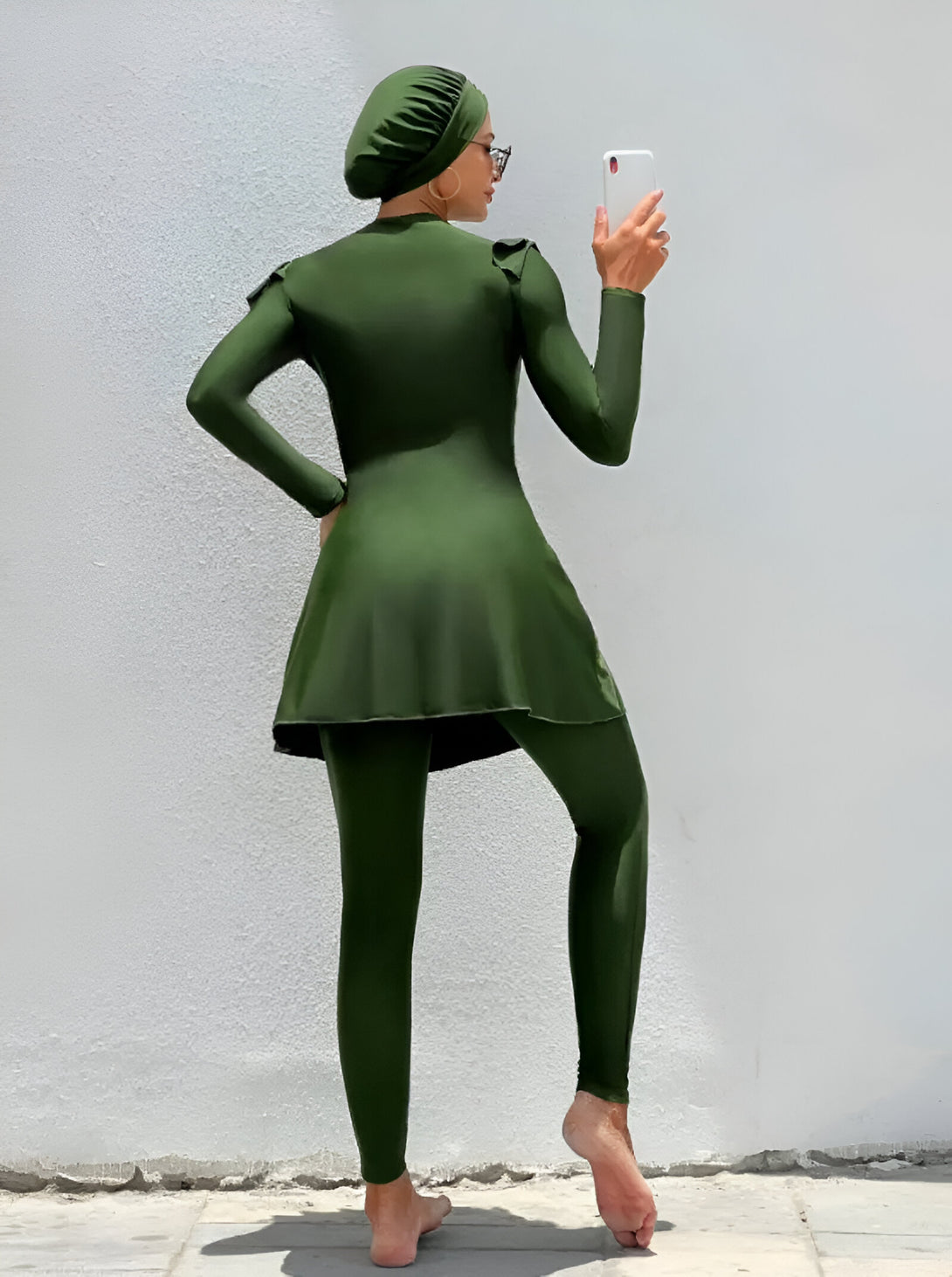 Une femme portant un maillot de bain musulman vert foncé à manches longues et un hijab assorti.  Le maillot de bain est composé d'une robe longue et d'un pantalon.  Elle se tient devant un mur blanc.