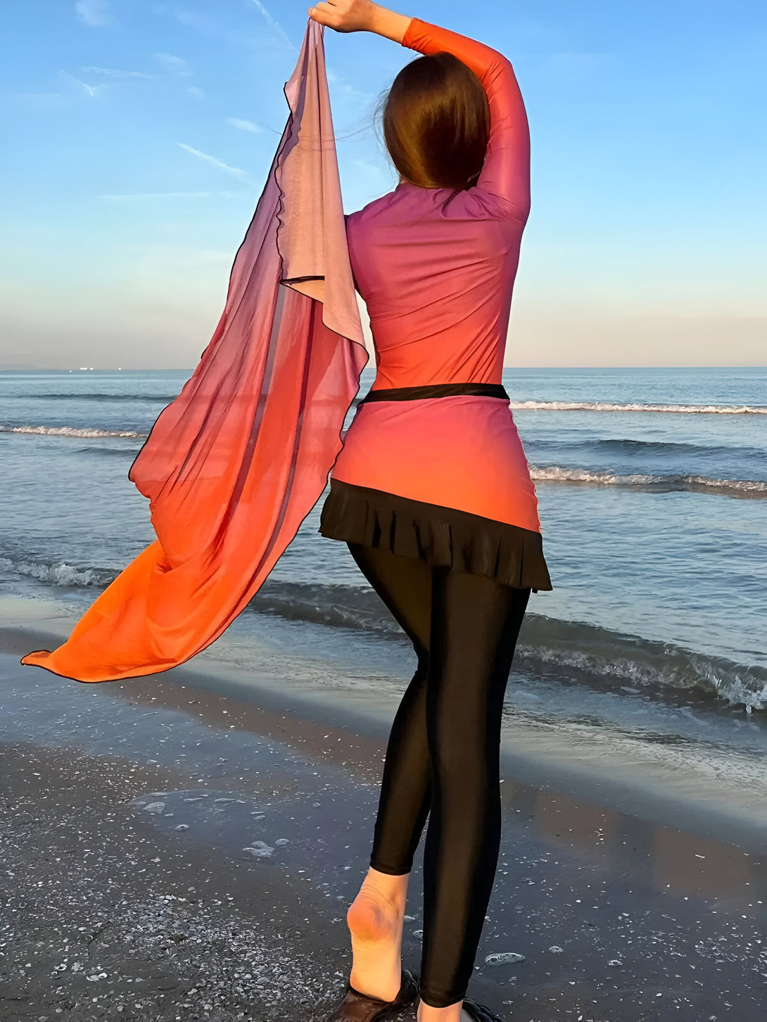 Une femme portant un hijab et un maillot de bain islamique sur une plage.  Le maillot de bain est de couleur rouge orangé avec une jupe noire. La femme porte un hijab orange et elle se tient pieds nus sur le sable.