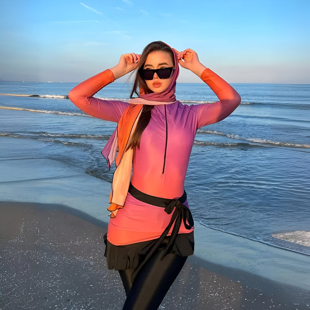 Une femme portant un maillot de bain musulman rose et orange avec un foulard assorti se tient sur une plage, portant des lunettes de soleil.
