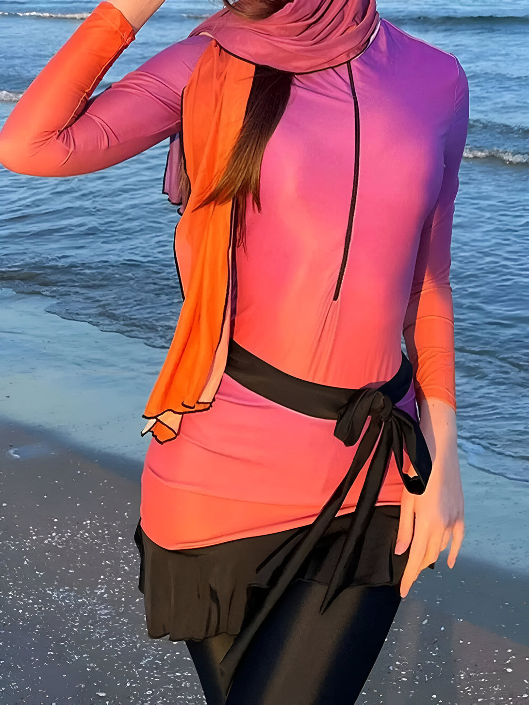 Une femme portant une tenue de plage musulmane élégante avec un chemisier à manches longues et un jupe en couleur dégradé orangée et violette.  La jupe est attachée avec une ceinture noire et elle porte un hijab orange.  Le modèle est sur une plage de sable.