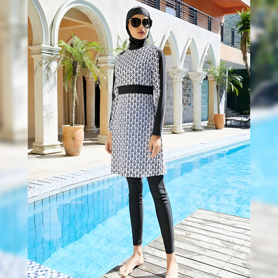 Une femme portant un maillot de bain musulman noir et blanc à motifs floraux se tient près d'une piscine. Elle porte également un hijab noir et des lunettes de soleil.