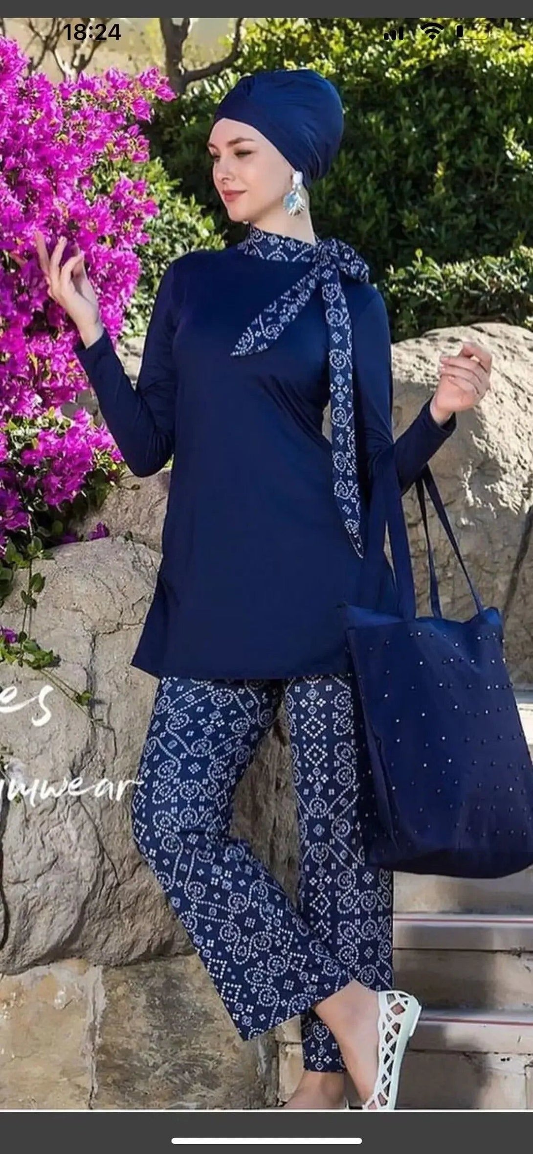 Une femme portant un hijab bleu marine et un maillot de bain musulman bleu marine à motifs blancs. Elle porte un sac à main bleu marine et des sandales blanches.