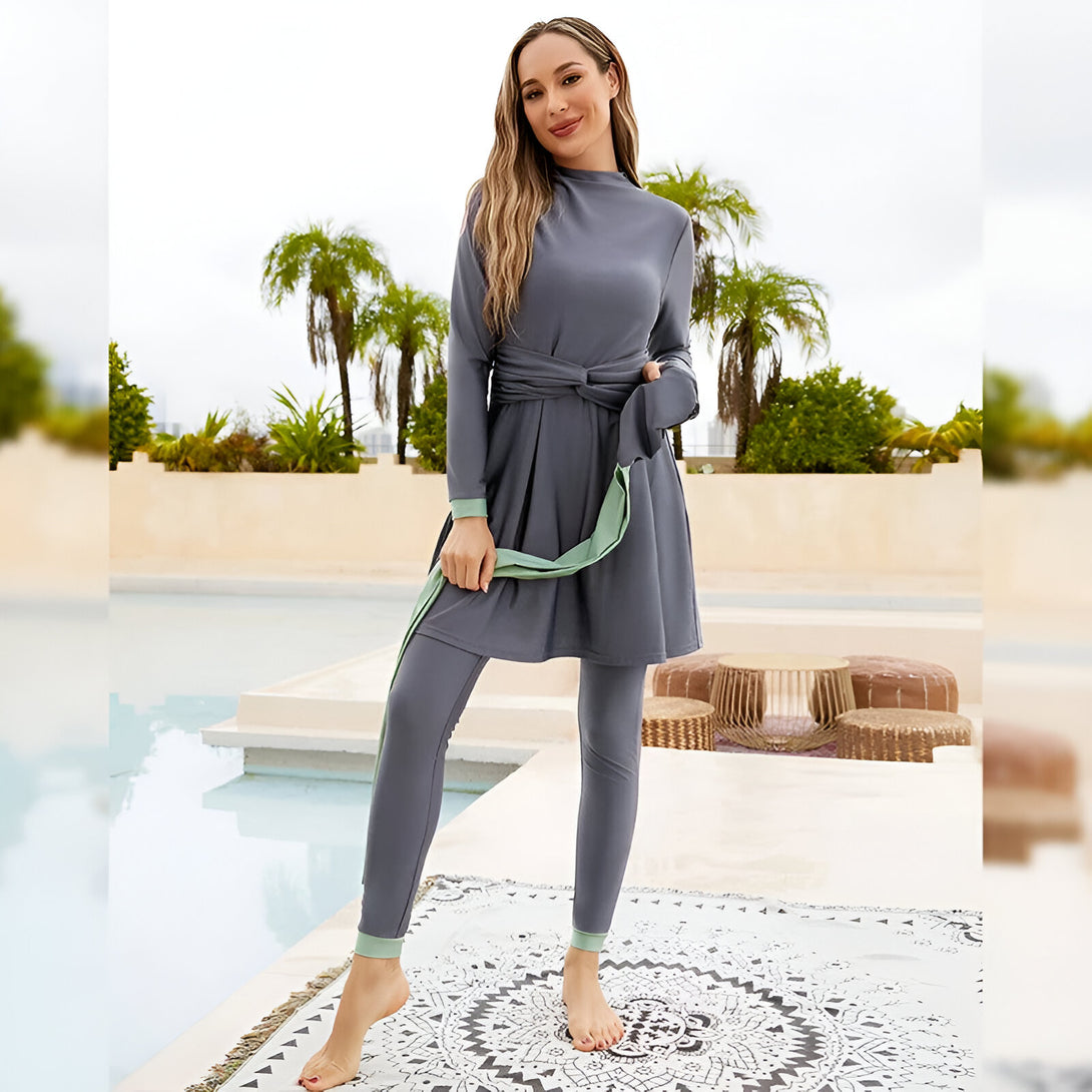 Une femme portant un maillot de bain musulman gris à manches longues et un pantalon assorti. Le maillot de bain a une ceinture à nouer à la taille et des détails en vert menthe. La femme est debout devant une piscine.