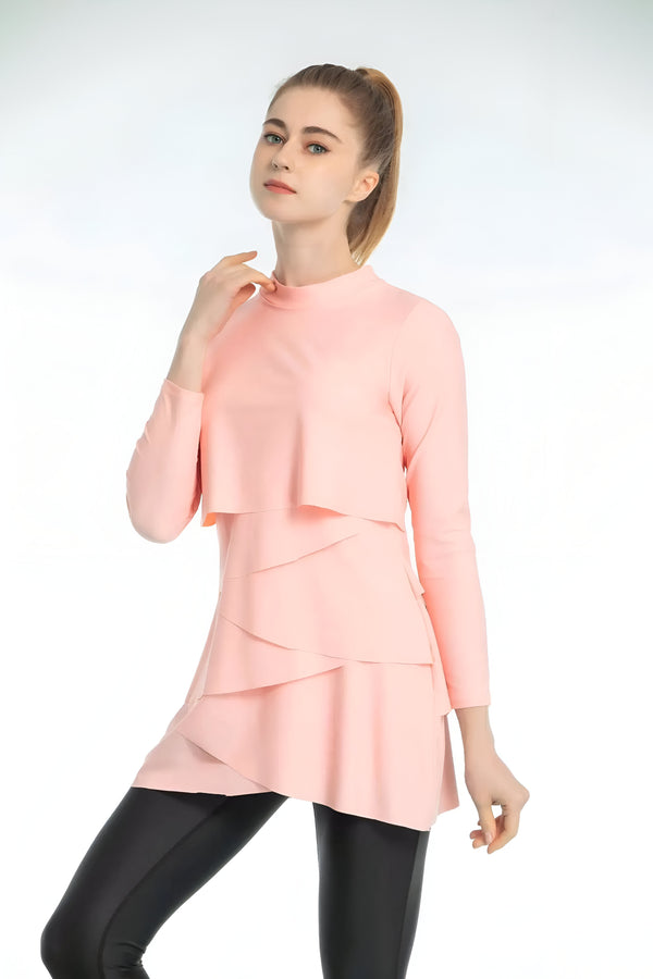 Une femme porte une tunique rose à manches longues avec des volants. La tunique est cintrée à la taille et tombe au-dessus des genoux. Elle porte également un pantalon noir.
