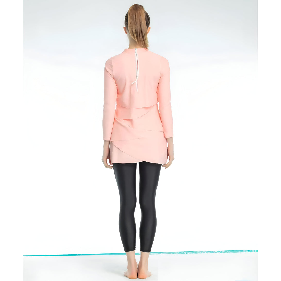 Une femme porte une robe rose à manches longues avec une fermeture éclair dans le dos et un pantalon noir. La robe est à couches multiples et le pantalon est ajusté. La femme est debout sur un fond blanc et a les cheveux tirés en arrière.  #habillementmusulman #tenueislamique #modemodeste #robe #pantalon #rose #noir