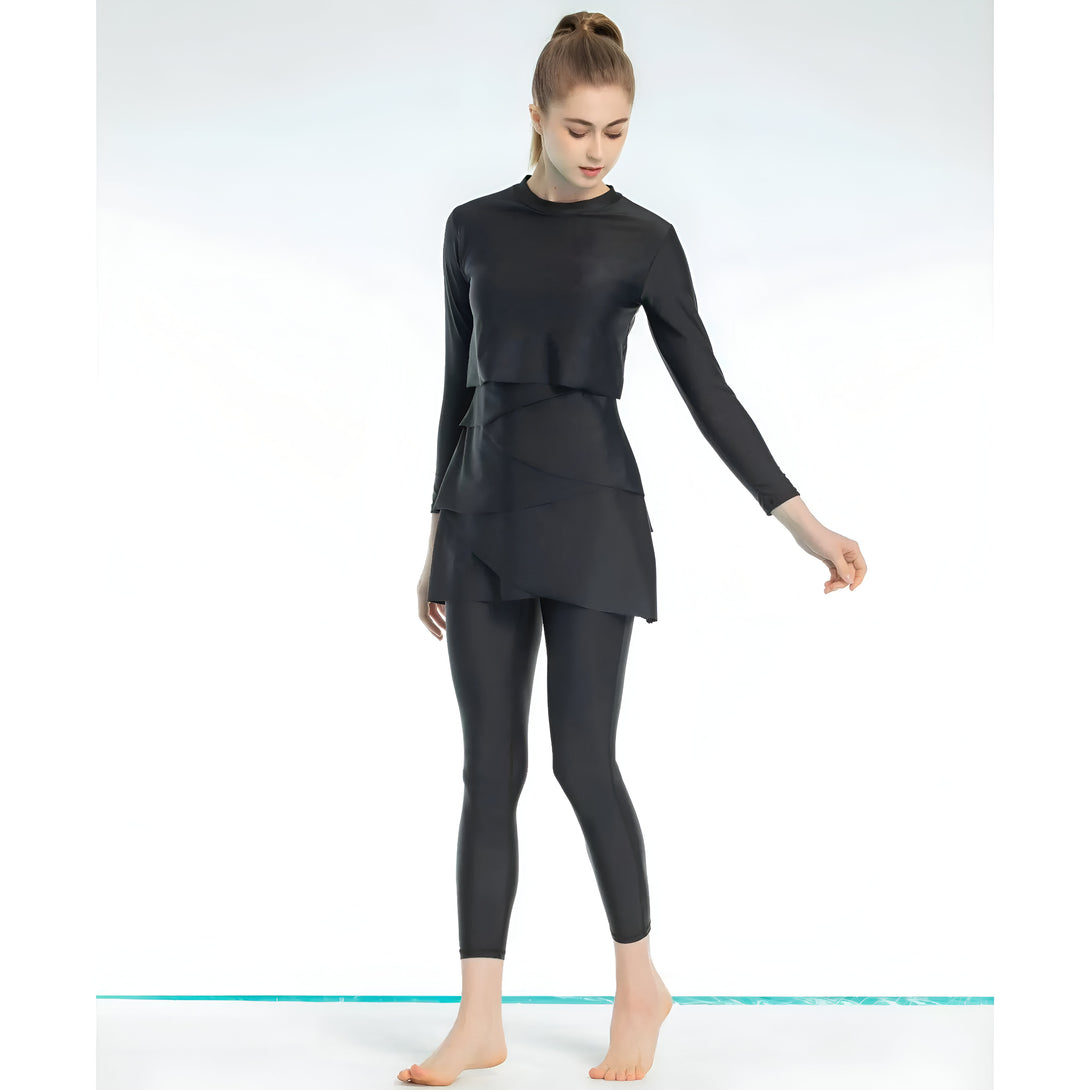 Une femme portant un maillot de bain musulman noir avec une jupe superposée. Le maillot de bain est à manches longues et couvre tout le corps. La femme est debout sur un fond blanc.