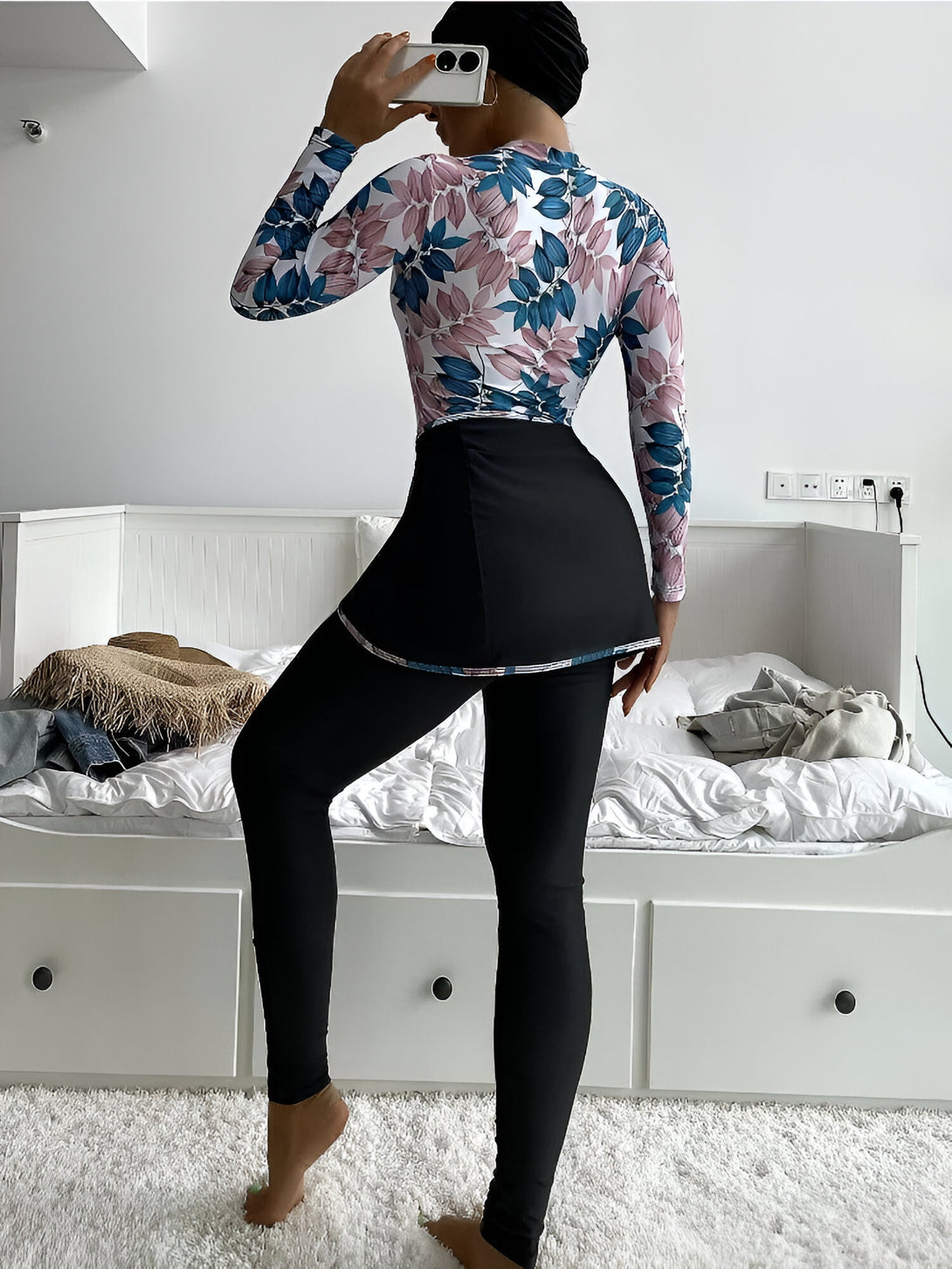 Une femme portant une tenue de bain musulmane à manches longues avec un motif floral. Le maillot de bain est blanc avec des feuilles bleues et roses. La jupe est noire et la tenue est complétée par un pantalon noir.