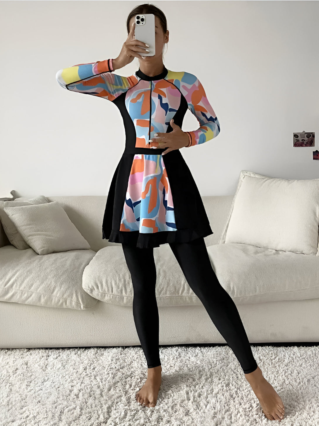 Une femme portant une robe de bain musulmane à manches longues avec un motif coloré et une jupe. La robe de bain est noire avec des détails colorés. La femme est debout devant un canapé blanc.