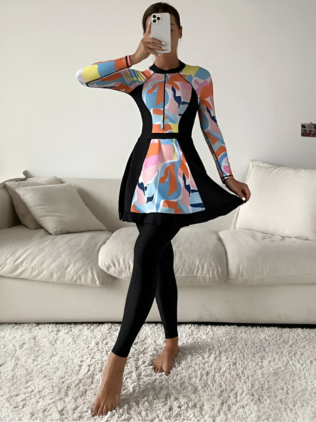 Une femme porte une robe de bain musulmane à manches longues avec un motif abstrait coloré et un pantalon noir. La robe a une jupe qui tombe juste au-dessus des genoux. Elle est debout sur un tapis blanc moelleux devant un canapé blanc.